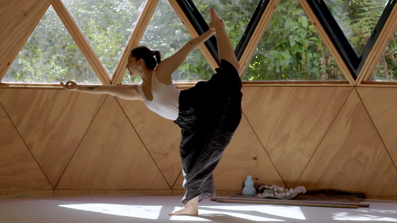 Junge Frau praktiziert Yoga in einem geometrischen Freiluft-Studio, konzentriert sich auf friedliche Bewegungen, steht auf und streckt sich nach vorne und außen, während sie ein Bein hält.