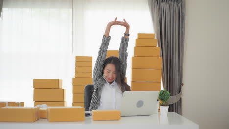 a young asian woman surrounded by products to be shipped stretches after working long hours on her laptop