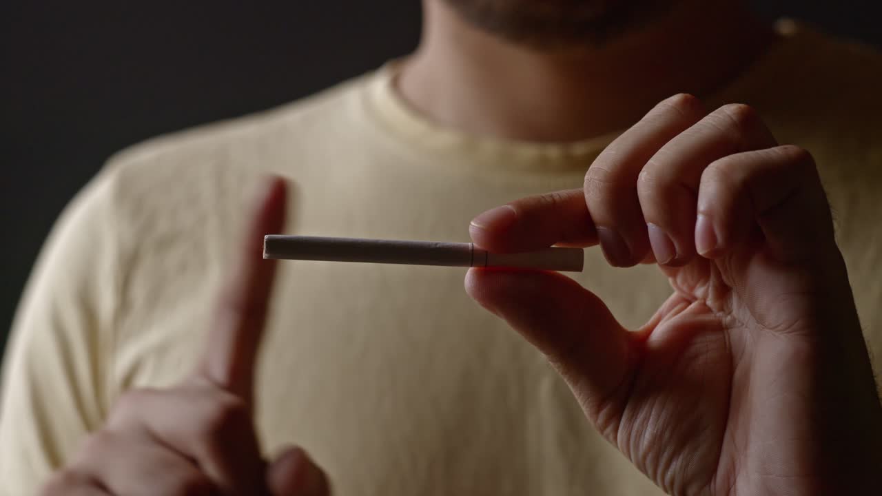 Close up of man in yellow shirt holding cigarette with one hand and waving index finger with the other to signal disapproval, dark background