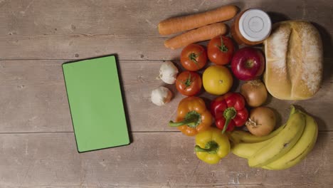 Overhead-Studio-Shot-Of-Hand-Picking-Up-Basic-Fresh-Food-Items-With-Green-Screen-Digital-Tablet-On-Wooden-Surface-1