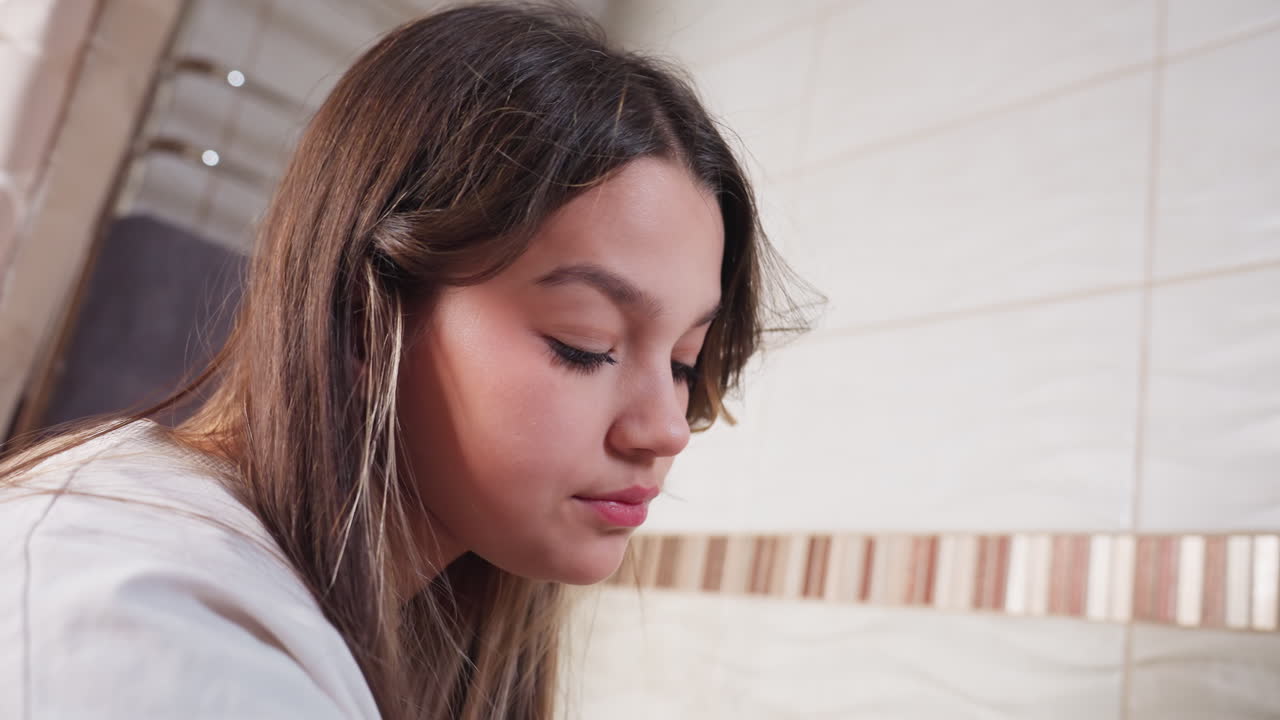 Beautiful young woman seated in bathtub, side profile closeup, eyes focused downward while washing feet with handheld shower, calm self care routine in bright tiled bathroom, soft light, relaxed mood