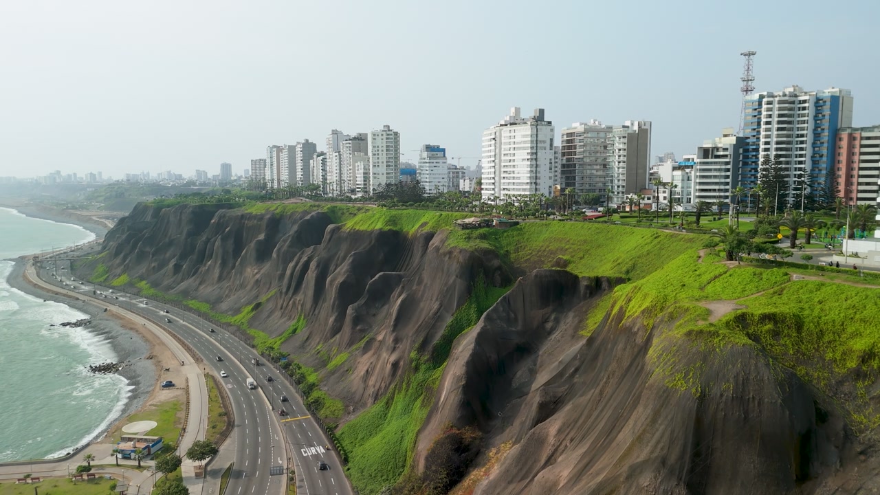 A slow rising aerial drone shot ascends along the dramatic cliffs of the Costa Verde in Lima, Peru, revealing the modern cityscape and the coastal highway below