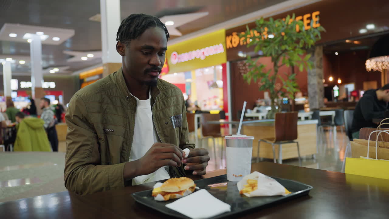 man wearing green jacket seated inside food court squeezing used napkin while looking around for bin to dispose, tray with half-eaten burger, fries, drink in front, restaurants visible in background