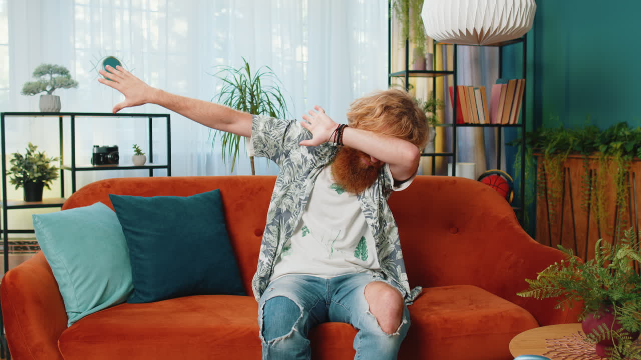 Happy man having fun, dancing, dabbing raising hands, making dub dance gesture at home room on sofa