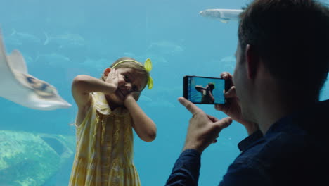 family at aquarium father using smartphone taking photo of daughter little girl making faces enjoying having fun at oceanarium