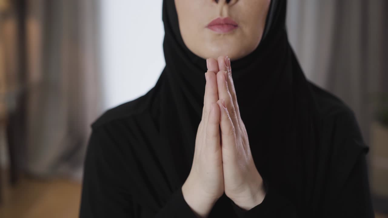 Close-up of unrecognizable Muslim woman folding hands together and praying. Young eastern lady talking with god at home. Religion, lifestyle, culture.