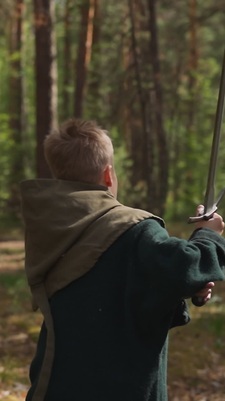 Kid in middle age costume with hood with sword runs in spring wood backside view. Young warrior of medieval period. Sports activity and education