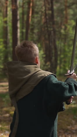 kid in middle age costume with hood with sword runs in spring wood backside view. young warrior of medieval period. sports activity and education