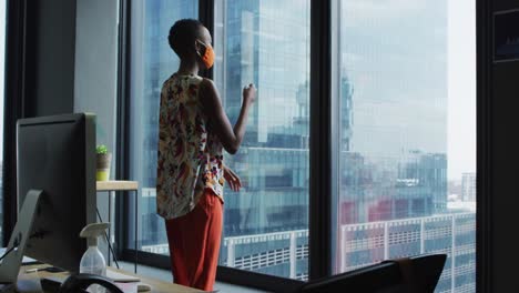 mujer afroamericana pensativa con máscara facial mirando por la ventana en una oficina moderna