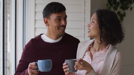 Romantisches-Hispanisches-Paar,-Das-Sich-Umarmt-Und-Im-Fenster-Steht-Und-Kaffee-Trinkt