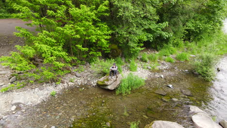 Person-sitting-on-a-rock-as-drone-flies-away-and-reveals-surrounding-area-of-the-Washougal-River-and-a-large-green-forest