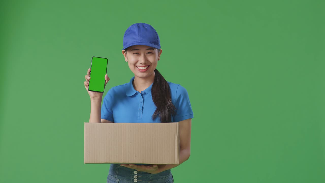 mensajera asiática en uniforme azul sonriendo y mostrando un teléfono inteligente de pantalla verde mientras entrega una caja de cartón en el fondo de la pantalla verde en el estudio