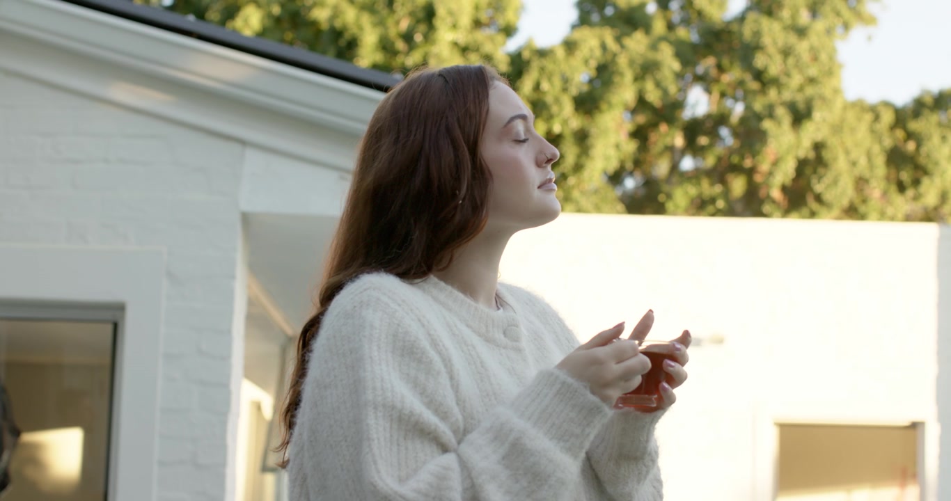 Donna con maglione bianco sulla soglia, con tazza rossa e cucchiaino, mescola prima di girarsi verso la luce del sole.