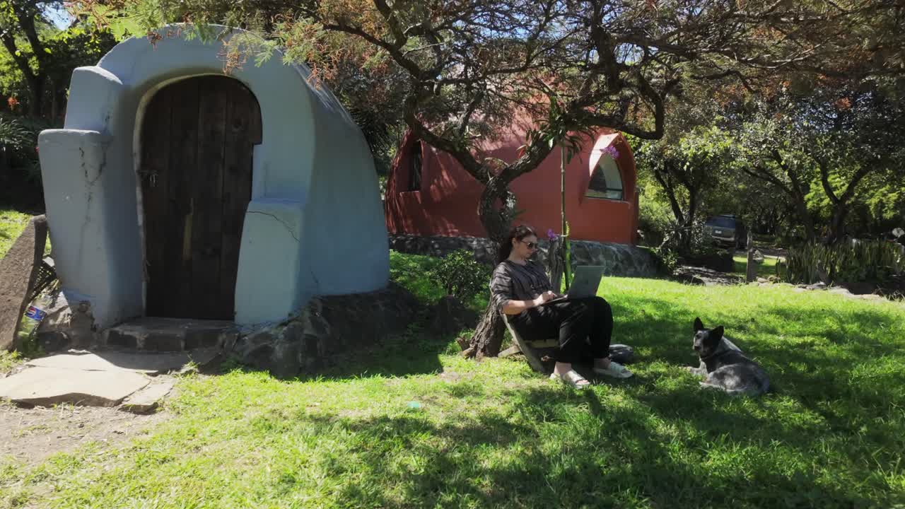 Person working on laptop under a tree with a dog beside dome-shaped eco-home