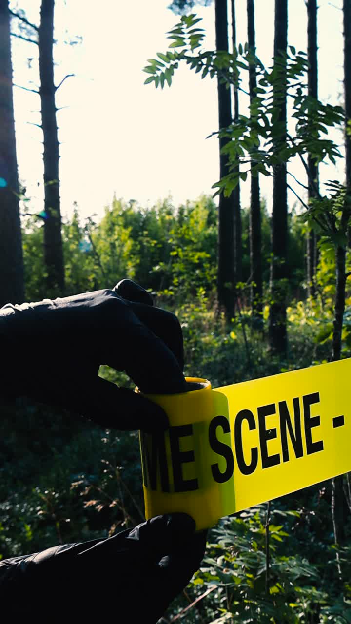 Vertical video of a yellow colored Crime Scene police caution tape ribbon being pulled and unraveled in front of a sunny forest by an investigator with black nylon gloves. Blurry bokeh background