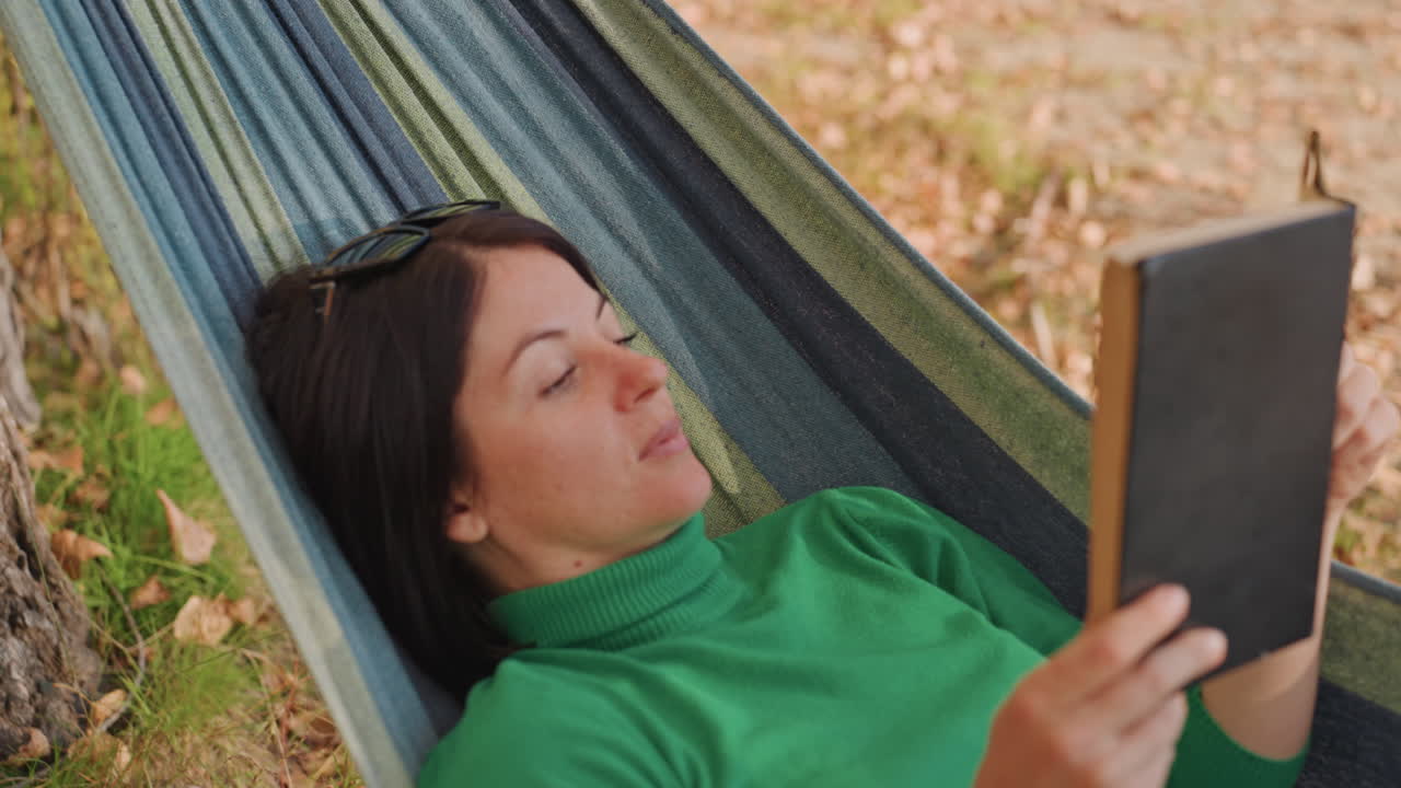 Donna che legge in amaca in campeggio sotto un albero, indossa un maglione verde e occhiali da sole. Foglie autunnali a terra, espressione calma, pagine che si girano lentamente. Un pigro ritiro pomeridiano per una fuga letteraria solitaria.