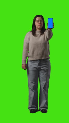 Vertical-Video-Full-Length-Shot-Of-Young-Woman-Holding-Blue-Screen-Mobile-Phone-To-Camera-Standing-Against-Green-Screen