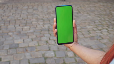 hand holding a smartphone with a green screen on cobblestone pavement