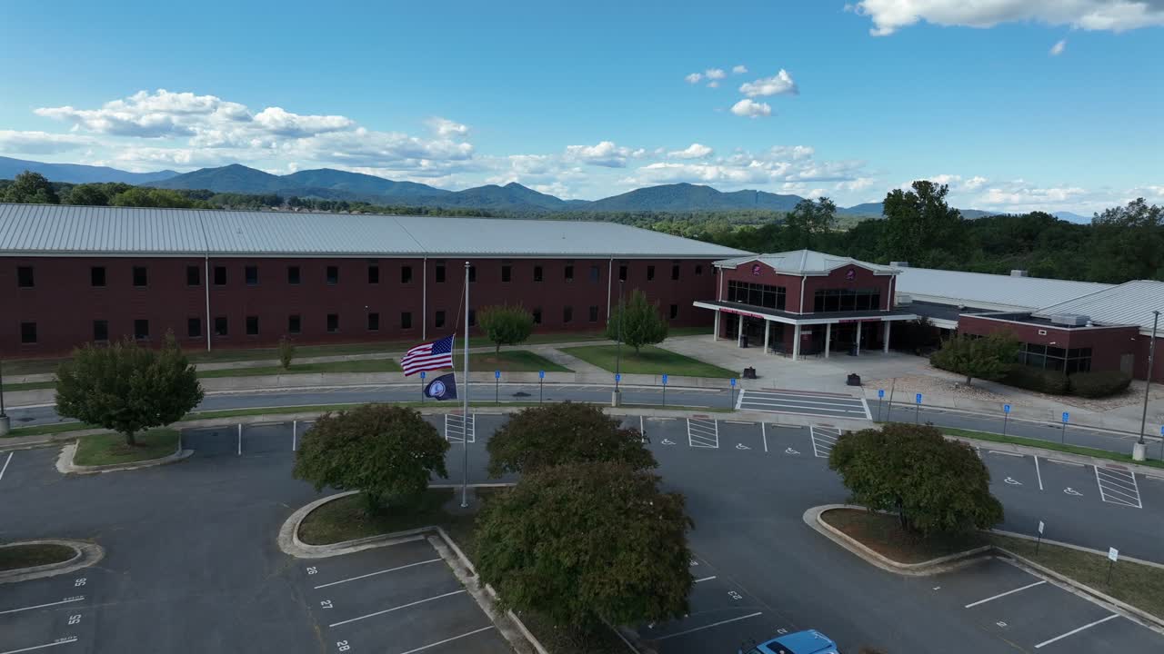 Aerial approaching shot of closed Jefferson Forest High School in Lynchburg with empty parking area. Waving flag of American and Virginia on flagpole. Patriotic city in USA
