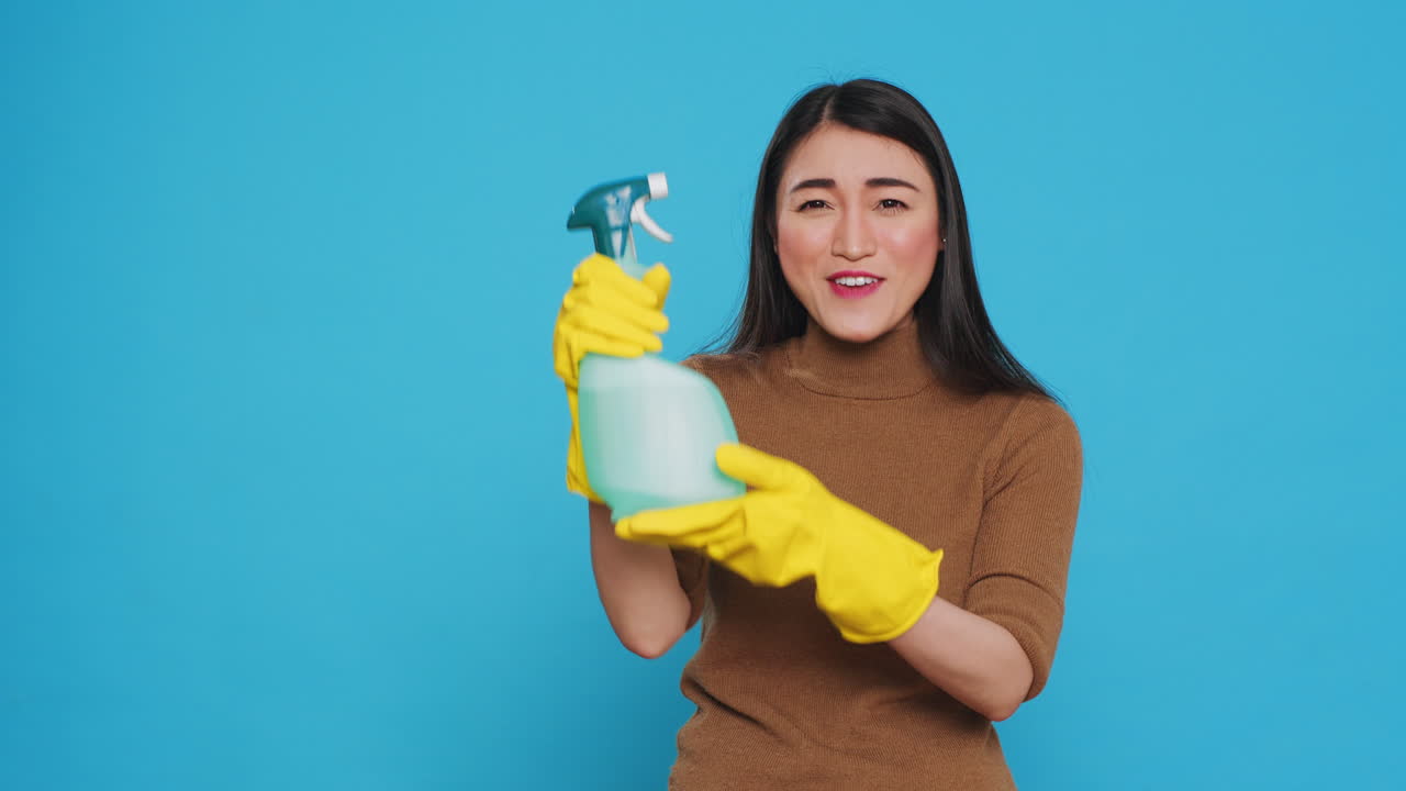 Smiling asian maid wearing protective gloves while showing chemical detergent spray in front of camera