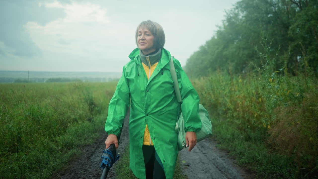 Woman in green raincoat carrying mint-colored bag and umbrella, walking along a muddy path surrounded by greenery and trees under a misty sky, looking into the distance