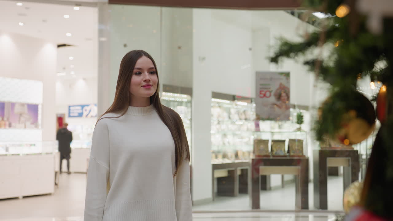 White girl walking towards decorative plant in bright shopping mall, with hair flowing down her back, blurred background shows another person walking, showcasing stylish and vibrant atmosphere
