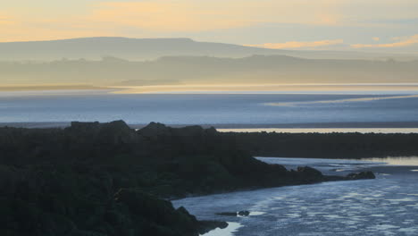 Rocky-shoreline-and-misty-landscape-beyond-bathed-in-early-morning-autumn-sunlight