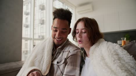 close up a happy young man with black skin a brunette with stubble in a checkered cream shirt sits on a modern sofa with his young girlfriend wrapped in a white woolen blanket. they read funny and happy stories to each other in a modern apartment