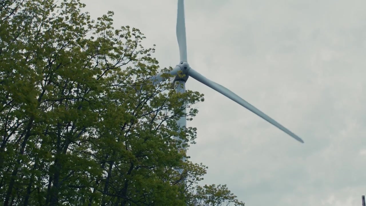 Carrellata di un mulino a vento che gira nel vento generando energia pulita dietro gli alberi in un campo, contro un cielo parzialmente nuvoloso, con uno stile e un movimento fluido e cinematografico.