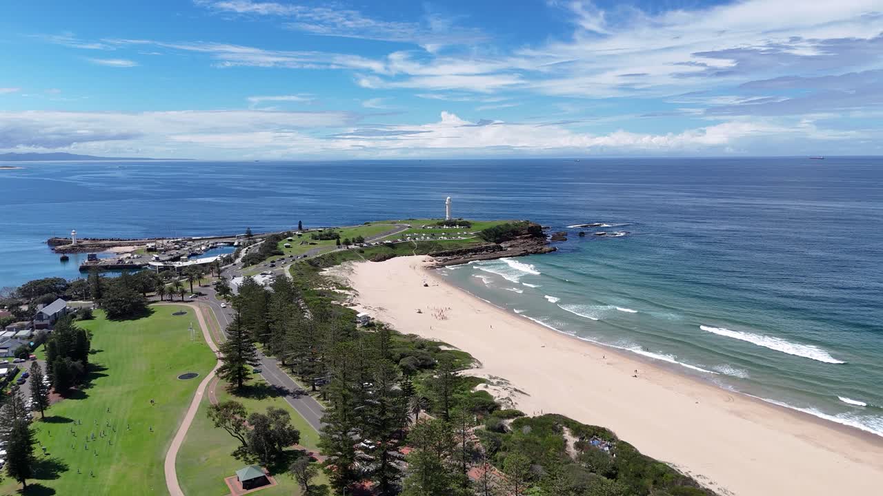 Wollongong foreshore and parkland stretch along beach with surf and walking paths visible, aerial establishing to monument lighthouse