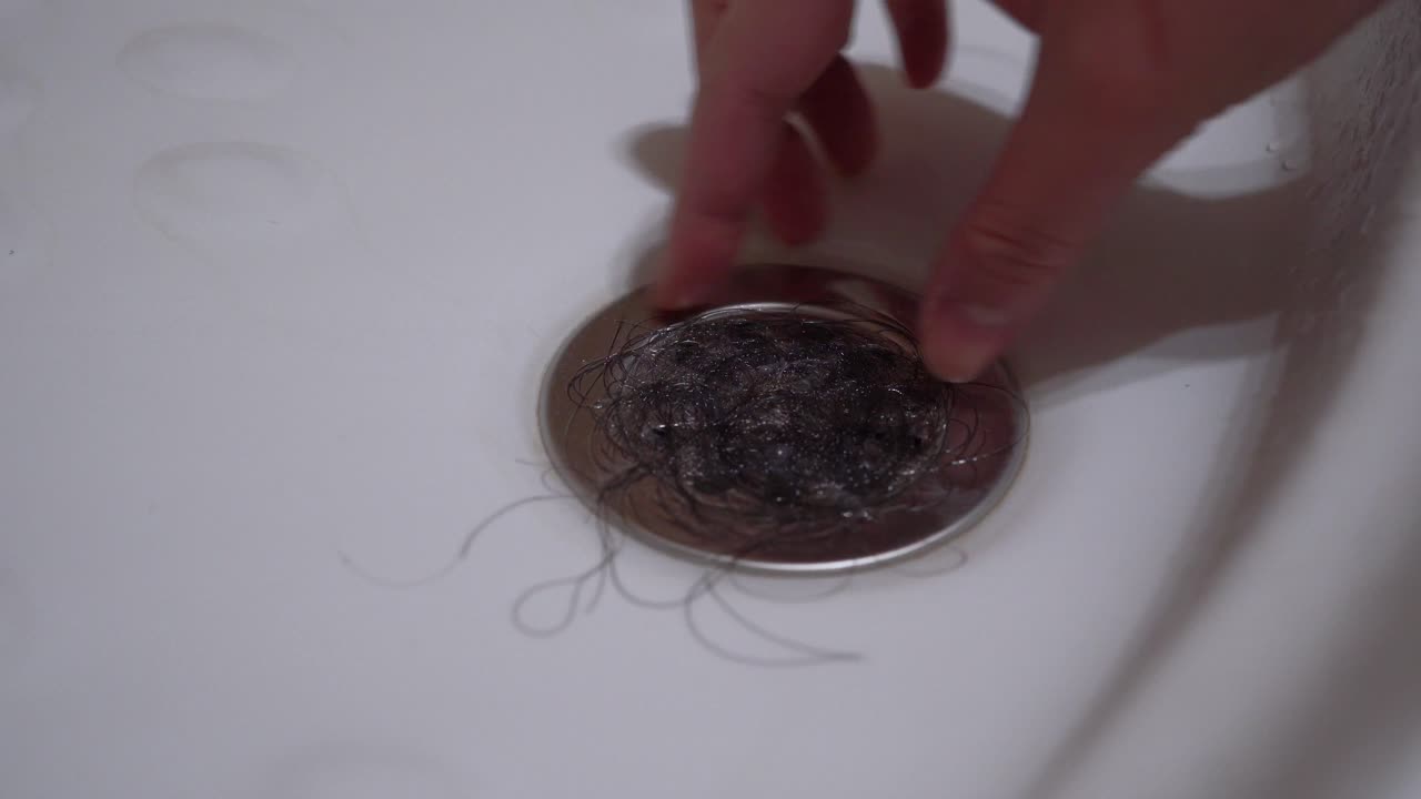 A female hand picks up black hair from the drain hole in the shower