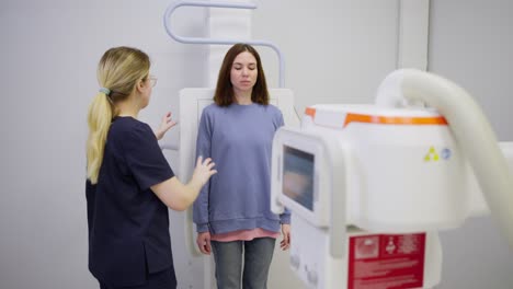 A-confident-brunette-girl-in-a-blue-jacket-stands-near-a-special-fluorography-installation-and-does-fluorography-under-the-supervision-of-a-girl-doctor-nurse-in-a-modern-clinic-in-a-bright-office