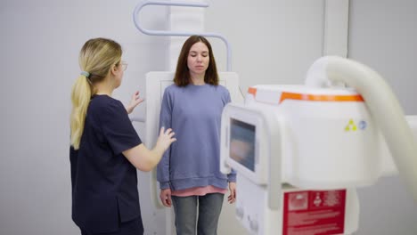 una chica morena confiada en una chaqueta azul se encuentra cerca de una instalación especial de fluorografía y hace fluorografía bajo la supervisión de una enfermera médica chica en una clínica moderna en una oficina brillante