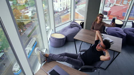relaxing break from work in the modern office downtown. man is half-lying in the chair and woman taking selfie. high angle view. city panorama at backdrop.