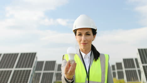 Retrato-De-Una-Ingeniera-En-Uniforme-Con-Casco-Protector-Que-Muestra-Una-Bombilla-A-La-Cámara-En-Una-Plantación-Solar