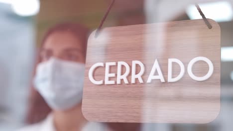 girl with face mask changing closed to open sign in spanish on window looking outside waiting for clients. start-up at covid time concept.