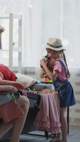 la niña ayuda a la madre a recoger artículos para el viaje en el sofá en la sala de estar. la mujer dobla la ropa y la niña con un sombrero elegante pone los juguetes en la maleta