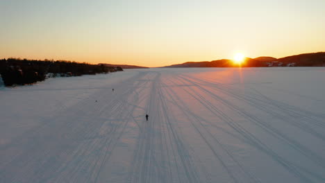 Vista-Aérea-De-Personas-Caminando-Con-Raquetas-De-Nieve-Y-Perros-Corriendo-A-Lo-Largo-De-Un-Río-Congelado-Mientras-Se-Pone-El-Sol