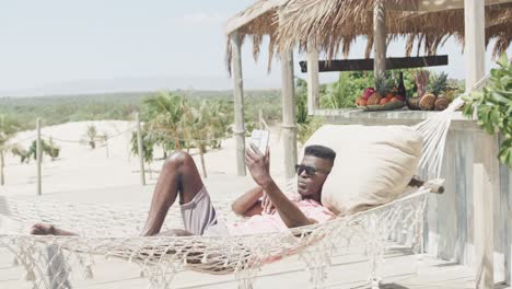 happy african american man lying in hammock taking selfie with phone on sunny beach, slow motion