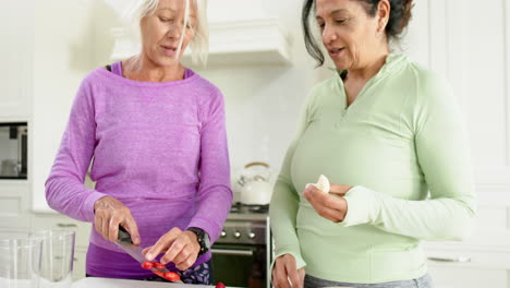 Two-happy-diverse-senior-women-chopping-fruits,-discussing-and-smiling-in-sunny-kitchen,-slow-motion