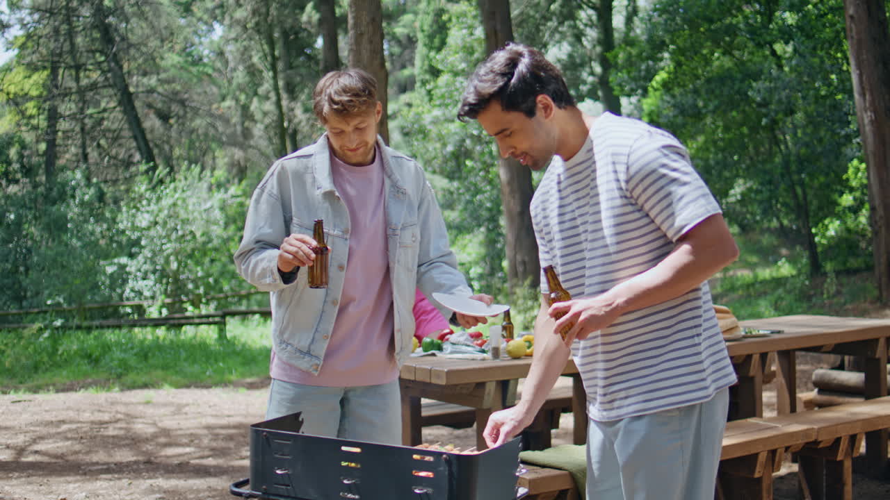Two men grilling food holding beer at summer forest barbecue closeup. Friends