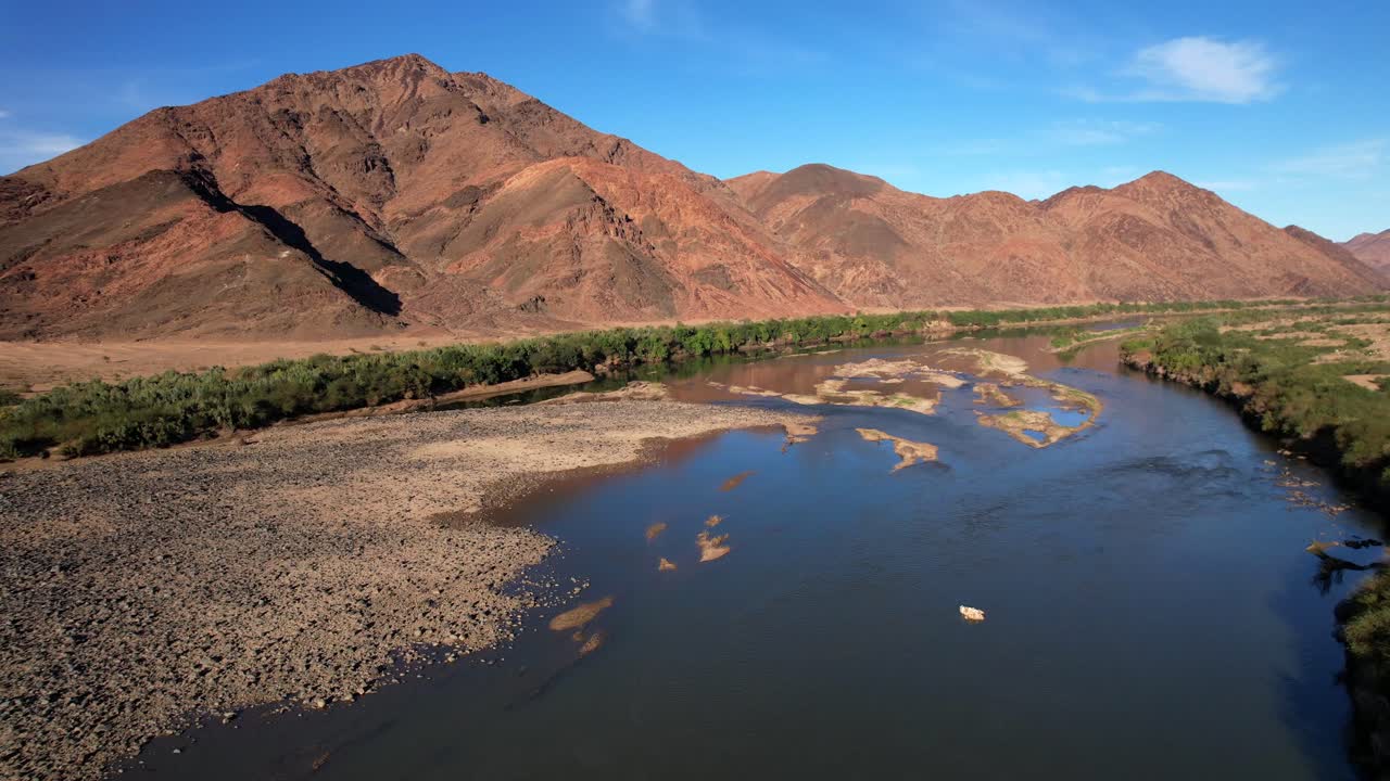 Drone ascends over a wide river flanked by dry rocky mountains and green riverbanks under a clear blue sky, showcasing vibrant nature and scenic landscape contrast