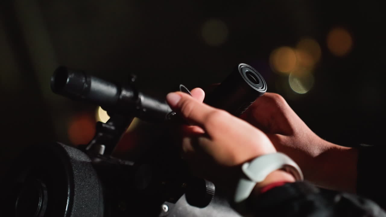 closeup hands mounting eyepiece on telescope, fingers fitting lens cap and rotating focus, small tools and wristwatch visible, careful teamwork and precise calibration against blurred city lights