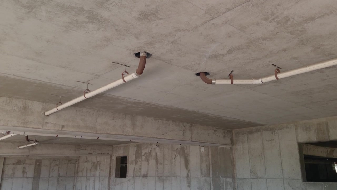 Steady pan revealing exposed ceiling pipes and unfinished concrete interior inside vacant structure.