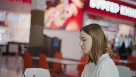 white chic woman seated reading a book with a coffee cup and shopping bag beside her, soft lighting and blurred background featuring other shoppers in a mall environment