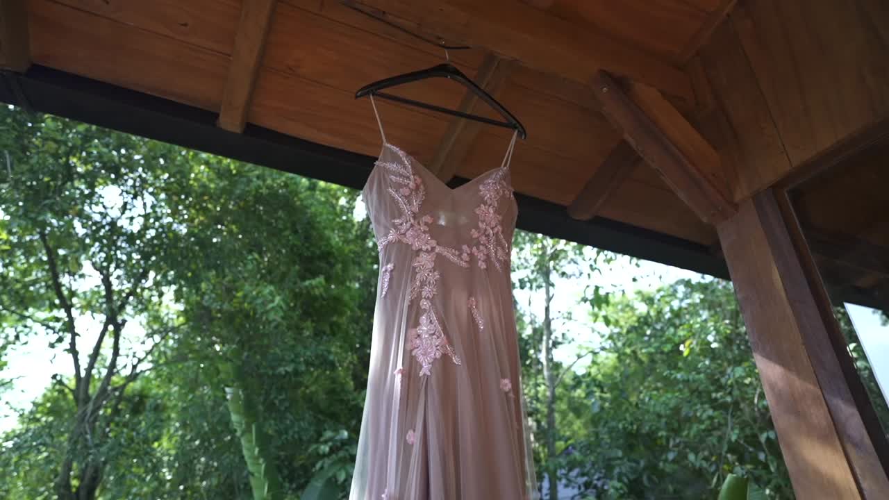 Rallentamento graduale che svela un bellissimo abito da sposa appeso a una gruccia con luce naturale e sfondo di fogliame da giardino durante la preparazione del matrimonio