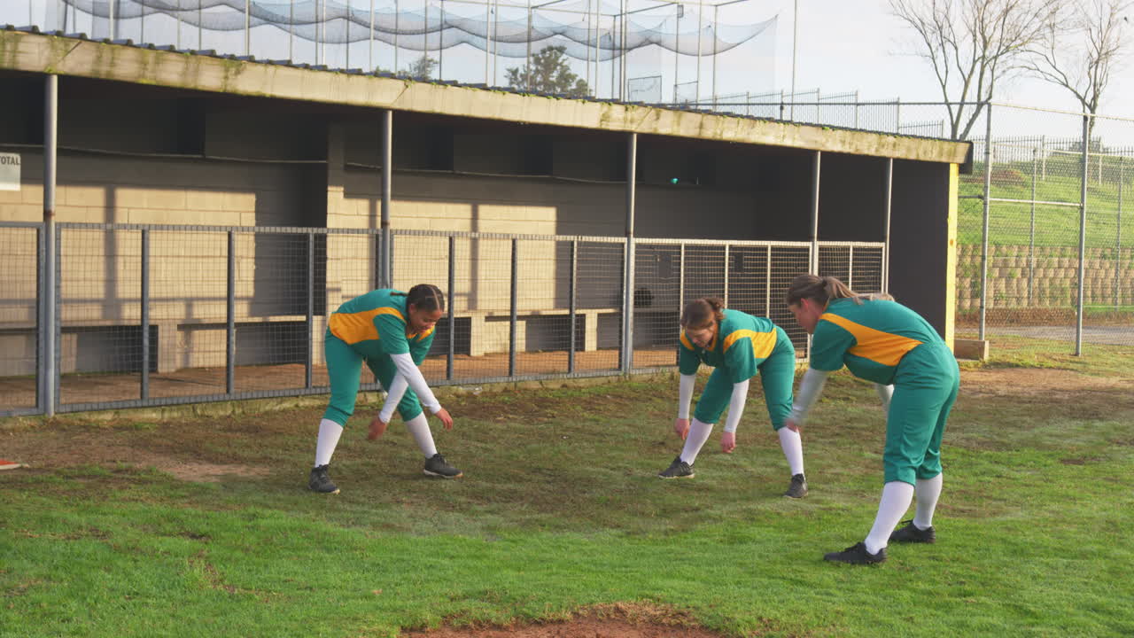 Multiracial female baseball players warming up, shaking arms and stretching legs on a pitch