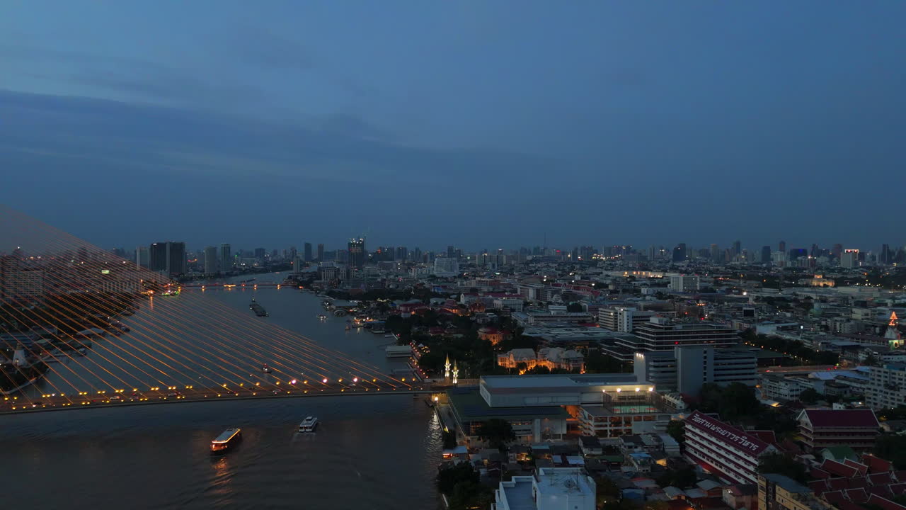 Aerial dusk view of Khin Lom Chom Saphan riverside restaurant under Rama VIII Bridge in Bangkok, with bridge lighting, river activity, and vibrant urban energy