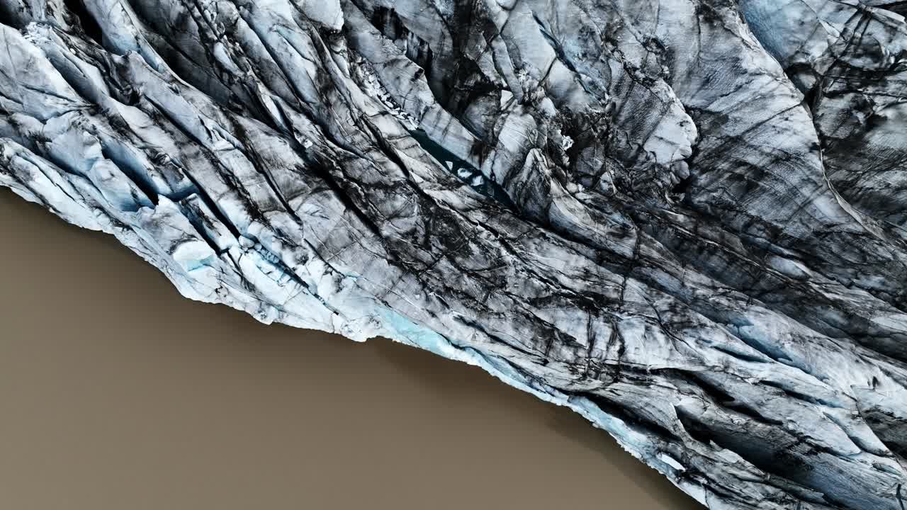 Aerial shot moving along a rugged Icelandic glacier edge where dark sediment lines cut through blue ice above a muddy glacial lake, revealing powerful natural contrast and raw northern energy