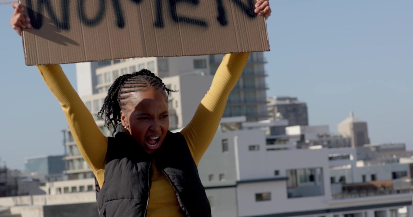 Donna afroamericana cammina su un tetto, si ferma vicino a una ringhiera, inclina un cartello con scritto 'DONNE' e protesta.